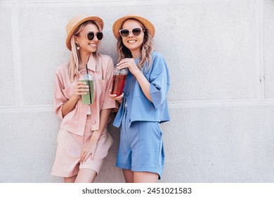 Dve mladé krásne usmievavé bedrové ženy v módnom letnom oblečení. Bezstarostné ženy pózujúce vonku. Pozitívne modelky držiace a pijúce čerstvé koktailové smoothie v plastovom pohári so slamkou