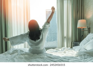 Hotel relaxation on lazy day with Asian woman waking up from good sleep on bed in weekend morning resting in comfort bedroom looking toward city view, having happy, work-life quality balance lifestyle