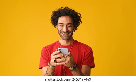 Uśmiechnięty mężczyzna z kręconymi włosami, ubrany w czerwoną koszulkę, korzystający z telefonu komórkowego, piszący masaże na żółtym tle w studiu