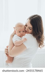 Una giovane mamma felice e un neonato sorridono, si abbracciano e si baciano su uno sfondo bianco isolato di una finestra: una famiglia amorevole a casa.