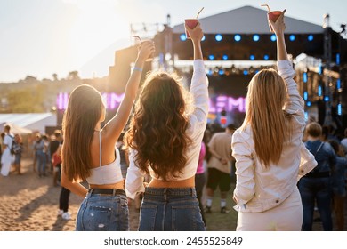 Vue arrière de trois jeunes femmes lors d'un festival de musique sur la plage avec des boissons en l'air, profitant de l'ambiance du concert. Tenues d'été décontractées, éclairage du coucher de soleil, foule et scène en arrière-plan, les filles font la fête ensemble en plein air.