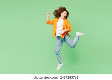 Full body young musician happy excited ginger woman she wear orange shirt white t-shirt casual clothes jump high playing air guitar isolated on plain pastel light green background. Lifestyle concept