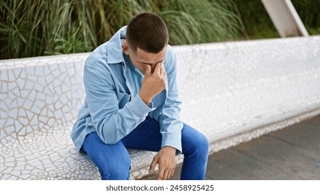 A young hispanic man in casual attire appears stressed while sitting on a park bench outdoors.