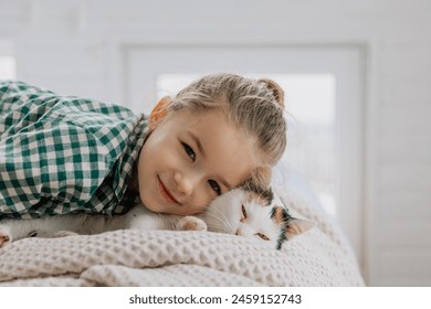 Seorang gadis kecil berambut pirang mengenakan piyama memeluk seekor kucing peliharaan di kamar tidurnya. Konsep pagi yang nyaman di rumah