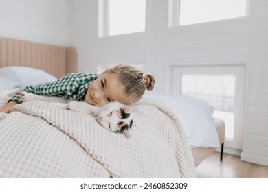 Seorang gadis kecil berambut pirang mengenakan piyama memeluk seekor kucing peliharaan di kamar tidurnya. Konsep pagi yang nyaman di rumah