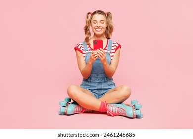 Gruaja e re me trup të plotë vesh pantallona të gjera xhins me bluza të kuqe, rroba të rastësishme blu, rrotulla blu duke lëvizur me rul ulur, përdorni celularin e izoluar në sfond të thjeshtë rozë Koncepti i stilit sportiv të verës për kohën e lirë
