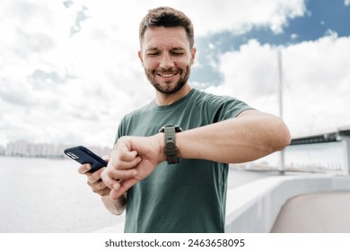 Egy férfi mosolyog, miközben az óráját nézi, és a kezében tartja a telefont, egy vízparton áll, híddal a háttérben.
