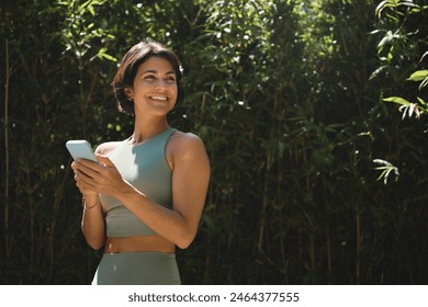 Jovem hispânica feliz em forma usando o telefone para verificar aplicativos de condicionamento físico, parada em um parque natural verde, olhando para longe, anunciando aulas de retiro de ioga ao ar livre, aplicativos de esporte online para celular. Espaço para texto.