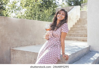 Beautiful young mixed race woman in pink dress smiling and holding glass of champagne. Attractive girl sits on stone parapet and drinks delicious wine outdoors. Granite steps and trees on background.