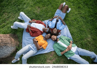 Group of thoughtful relaxed young African American friends lying together on green grass outdoors. Top view of four black students resting on plaid in park, enjoy nature and silence. Friendship.