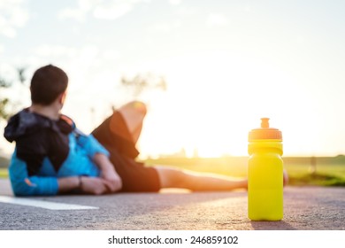 plastic sport bottle with water and blurred tired sportsman in shorts lying on the road in park and looking at sunset