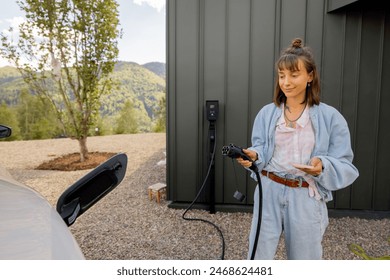 Mladá žena zapojuje nabíječku do elektrického vozidla poblíž jejího domu venku. Koncept moderních technologií, elektromobilů a udržitelného životního stylu