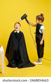 Cute little girl, acting as hairdresser, holds blowing hair of boy who loudly shouting against vibrant yellow background. Concept of children dreams, future career, occupation, games, fantasy. Ad
