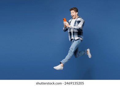 Całe ciało podekscytowane szczęśliwy młody człowiek nosi koszulę biały t-shirt swobodne ubrania skacze wysoko trzyma w ręku używa telefonu komórkowego odizolowanego na prostym niebieskim cyjanowym tle portret studyjny. Koncepcja stylu życia