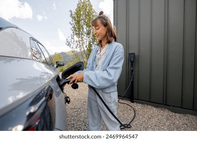Mladá žena zapojuje nabíječku do elektrického vozidla poblíž jejího domu venku. Koncept moderních technologií, elektromobilů a udržitelného životního stylu