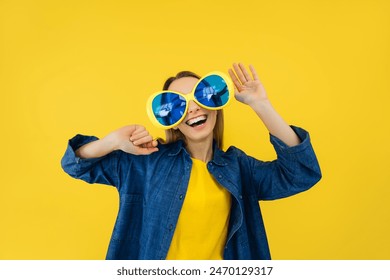 Young female wearing large big glasses and dancing while enjoying music over yellow studio background. Energetic woman in stylish clothes having great time while celebrating birthday.