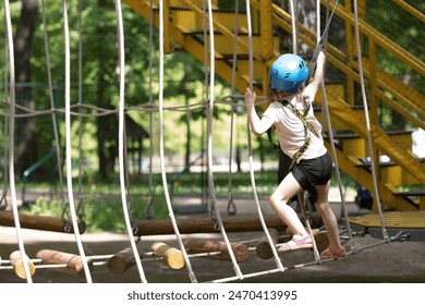 Summer. A small child is climbing a rope bridge in a rope park. A girl is having fun in an adventure park. A female kid on a climbing wall. Compliance with safety regulations.