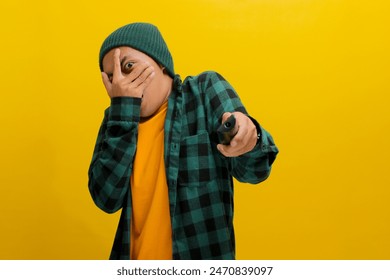 Un hombre asiático sorprendido, vestido con un gorro y una camisa informal, abre los ojos y la boca en estado de shock mientras ve una película de terror con escenas inesperadas e intensas, aislado sobre fondo amarillo