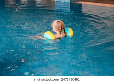 E klengt blondt Meedchen a Schwammmouwen schwëmmt am Pool. Sécher Schwammen, Sécherheet vun de Kanner um Waasser.