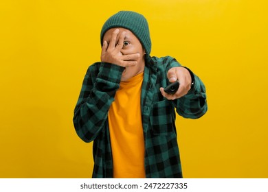 Un hombre asiático sorprendido, vestido con un gorro y una camisa informal, abre los ojos y la boca en estado de shock mientras ve una película de terror con escenas inesperadas e intensas, aislado sobre fondo amarillo