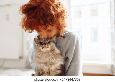 Gadis remaja berambut merah yang lucu duduk di lantai dekat jendela di rumah memeluk dan mencium kucing Maine Coon yang mendengkur. Persahabatan antara hewan peliharaan dan pemiliknya