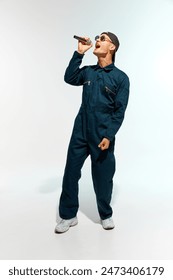 Artistic young man, musician dressed overalls with sunglasses singing with microphone against white studio background. Concept of music, festivals, contemporary art, hobby, creativity. Ad