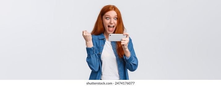 成就、目标和成功概念。快乐、快乐、可爱的红发少女握拳庆祝胜利，惊讶而满足地盯着智能手机显示屏，赢得游戏挑战。