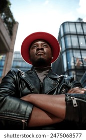Proud male model posing with arms crossed in front of modern buildings. He is wearing a red hat and black leather jacket. He is showing confidence and personality.