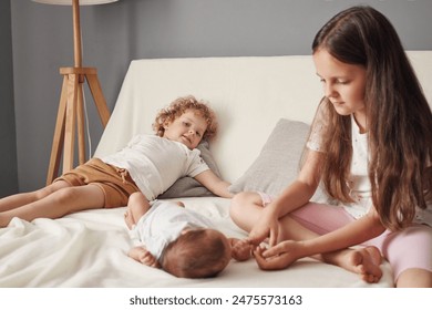 Sœurs aînées assises sur un lit dans une chambre lumineuse et confortable, petite sœur aux cheveux ondulés allongée sur un canapé tandis qu'une fille aux cheveux bruns joue avec un nouveau-né
