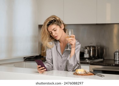 Young woman holding a glass of wine in her kitchen. Blond beautiful girl with phone drinking wine. Morning of pretty woman using social media on phone.