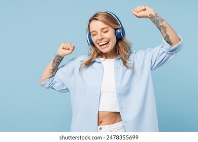 Young smiling happy woman she wears white top shirt casual clothes listen to music in headphones raise up hands dancing isolated on plain pastel light blue cyan background studio. Lifestyle concept