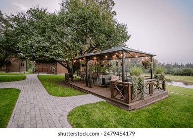 Blome, Lettonia - 11 settembre 2023 - Un accogliente patio esterno con luci a fili, piante sospese e comode sedute, situato su un rigoglioso prato verde con sentieri e circondato da alberi.
