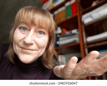 Ženska se nasmehne in iztegne roko na fotografiji od blizu. Ima blond lase, modre oči in nosi vijoličen pulover