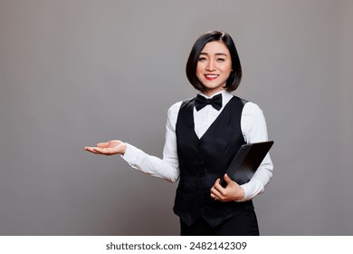 Garçonete asiática sorridente segurando tablet digital e dando boas-vindas ao hóspede com retrato de mão. Recepcionista de restaurante em pé com dispositivo portátil e mostrando com o braço enquanto olha para a câmera