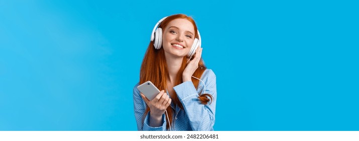 Technology, music and entertainment concept. Cheerful attractive redhead caucasian woman listening music in headphones, holding smartphone, smiling delighted, standing blue background.