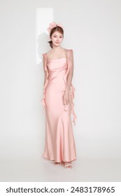 Elegantna mlada modna azijska ženska v osupljivi roza obleki, trendovsko naličena s svetlobo, ki sije skozi režo na njej, pozira na izoliranem belem ozadju.