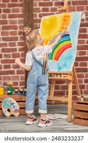 Bildung. Porträt eines fröhlichen kleinen Mädchens, mit bunten Farben befleckt und mit Pinseln in den Händen, vor einer Staffelei mit einer Kinderzeichnung. Kreativität der Kinder. Zeichnen und Fantasie der Kinder.