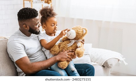 E jonkt afroamerikanescht Meedche sëtzt mat hirem Papp op enger Canapé. Si hält en groussen brongen Teddybier a weist op seng Nues, während hire Papp si ulächelt.