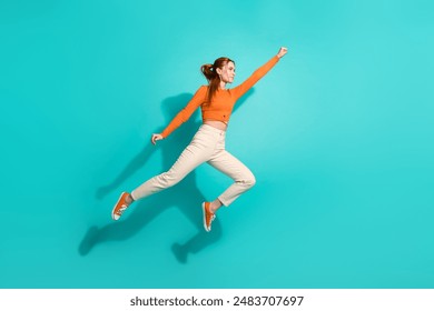 Foto van een sterke vrouw in volle lengte, die een crop top en een witte broek draagt ​​en haar vuist omhoog steekt en naar de lege ruimte vliegt, geïsoleerd op een turquoise achtergrond.
