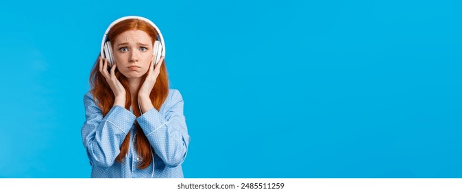 Girl about cry from emotional song. Upset redhead girlfriend suffering breakup, sobbing listening music in large white headphones, standing blue background in nightwear, unhappy.