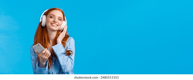 Technology, music and entertainment concept. Cheerful attractive redhead caucasian woman listening music in headphones, holding smartphone, smiling delighted, standing blue background.