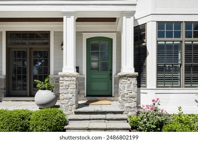 A front door detail on a white modern farmhouse home with a green front door and a covered porch with stone flooring and pillars. No brands or labels.