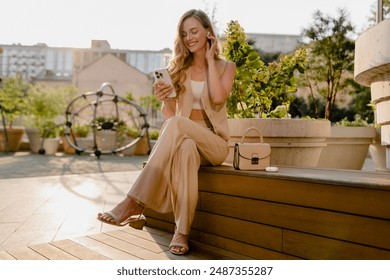 güneşli şehir caddesinde oturan güzel gülümseyen sarışın kadın, telefon, kentsel moda stili, yaz trend sezonu, iş kadını, göz alıcı saç modeli, yelek ve pantolon giyen bej takım elbise