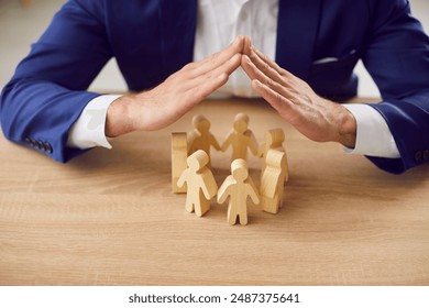 Geschäftsführer des Arbeitgebers schützt die Rechte der Arbeitnehmer. Mann hält Hände über einer Gruppe kleiner hölzerner menschlicher Puppenfiguren auf dem Tisch. Metapher für Sorgfalt, Schutz und Sicherheit am Arbeitsplatz