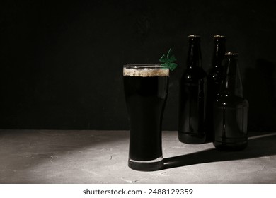 Copo e garrafas de cerveja escura com trevo para a celebração do Dia de São Patrício em uma mesa cinza grunge contra fundo preto