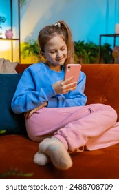 Young child girl texting share messages content on smartphone social media applications online, watching relax movie. Teenager kid uses mobile phone at home in evening room sitting on sofa. Vertical