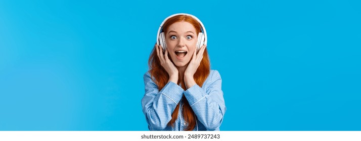 Excited and amused attractive caucasian redhead woman in pyjama, screaming from fascination and amazement hear new song, wearing headphones enjoy music, standing blue background.
