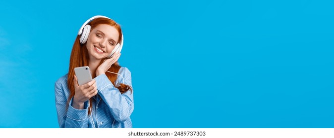 Tenderness, women and lifestyle concept. Cheerful tender redhead female in pyjama, tilting head enjoying listening music, holding smartphone and smiling as touching earphones from delight.