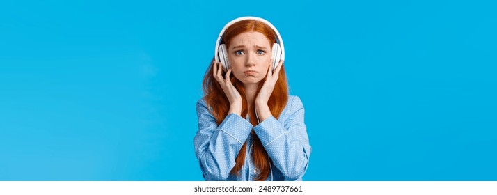 Girl about cry from emotional song. Upset redhead girlfriend suffering breakup, sobbing listening music in large white headphones, standing blue background in nightwear, unhappy.