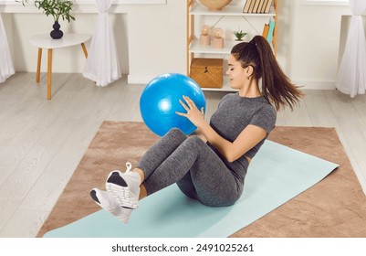 Gadis muda cantik melakukan latihan sambil berbaring di atas matras yoga dengan bola kebugaran di rumah. Gadis Kaukasia yang sporty melakukan gerakan russian twist menggunakan bola kebugaran. Konsep menjaga gaya hidup sehat.